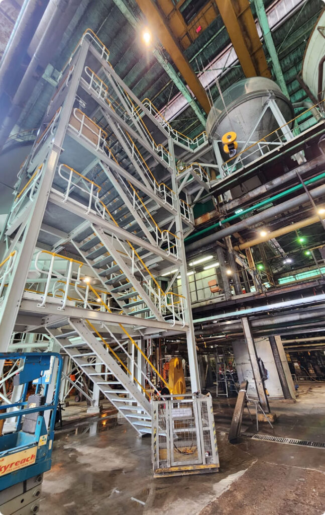 Complex industrial machinery with large pipes, metal structures, and flanges. The view is from below, showing the underside of a cylindrical vessel or tank connected to various pipes and support structures.